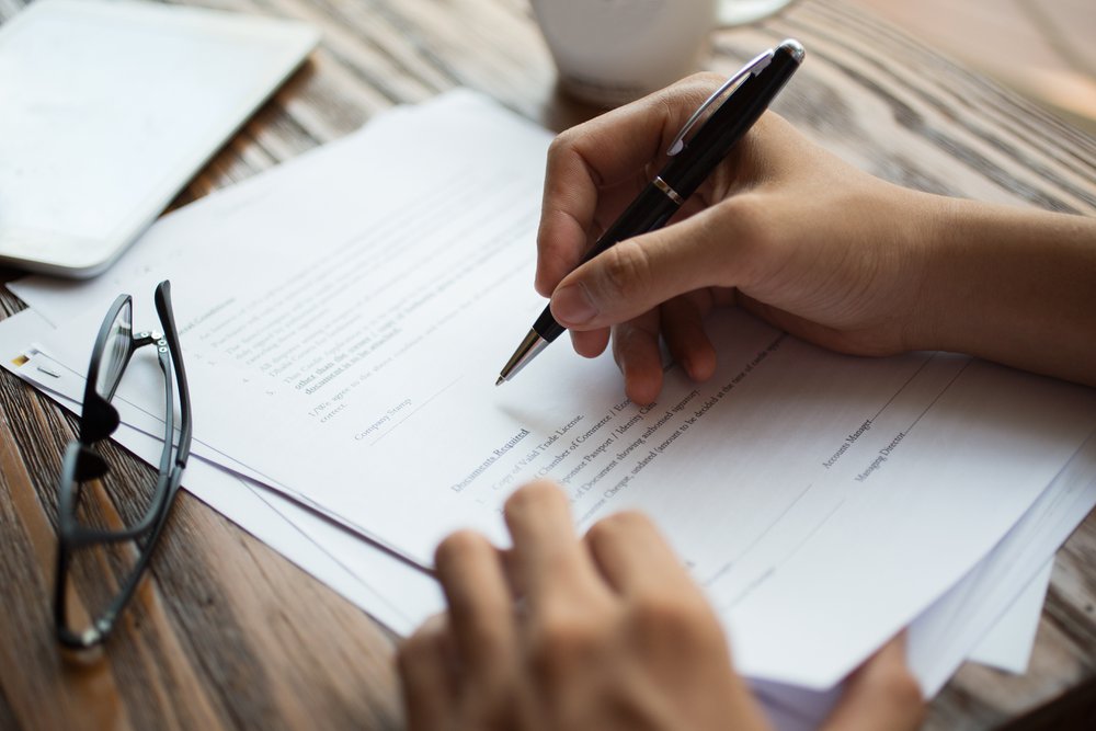 Unrecognizable businessman examining papers at table. Manager with ballpoint pen filling business papers. Close-up of male hands working at desk. Analyzing documents concept